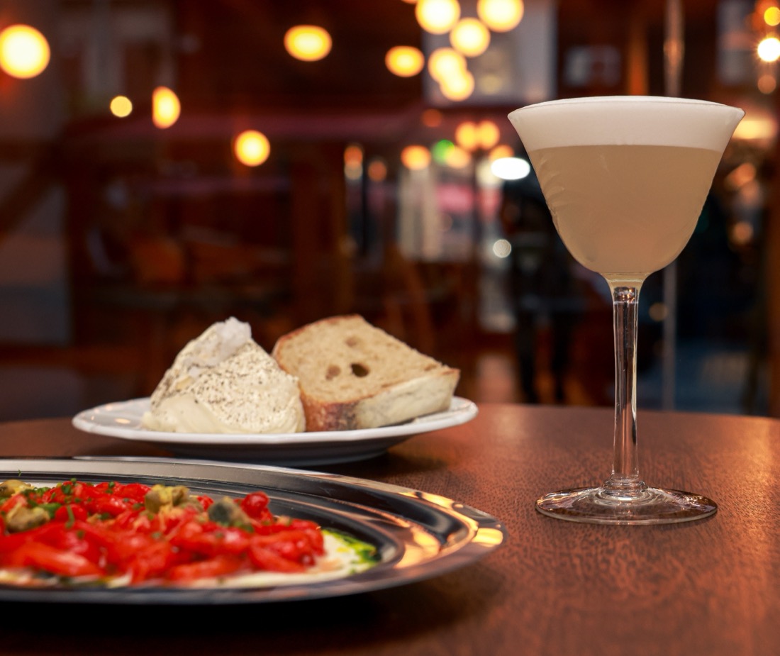 Un cóctel con espuma junto a pan y un plato de morrones asados, sobre una mesa de madera en Remy.